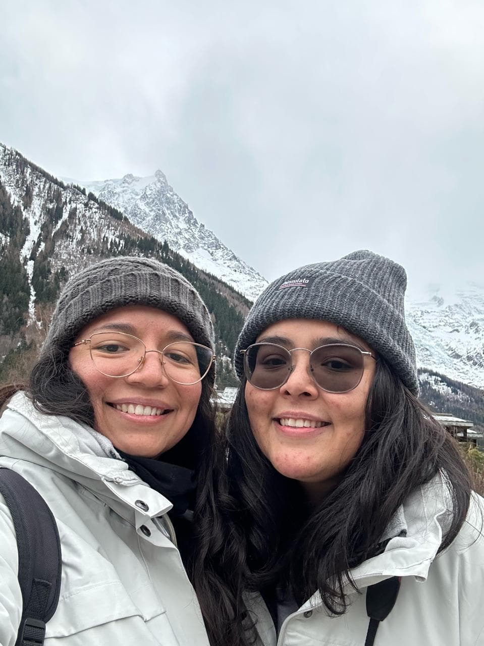 Mariel y Ale sonriendo juntas con gorros de invierno frente a un paisaje nevado de montaña.